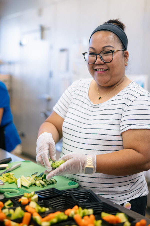 Chef Genie sorts sliced vegetables into takeout containers. She wears a white, striped shirt and glasses. Her hair is tied back into a bun.