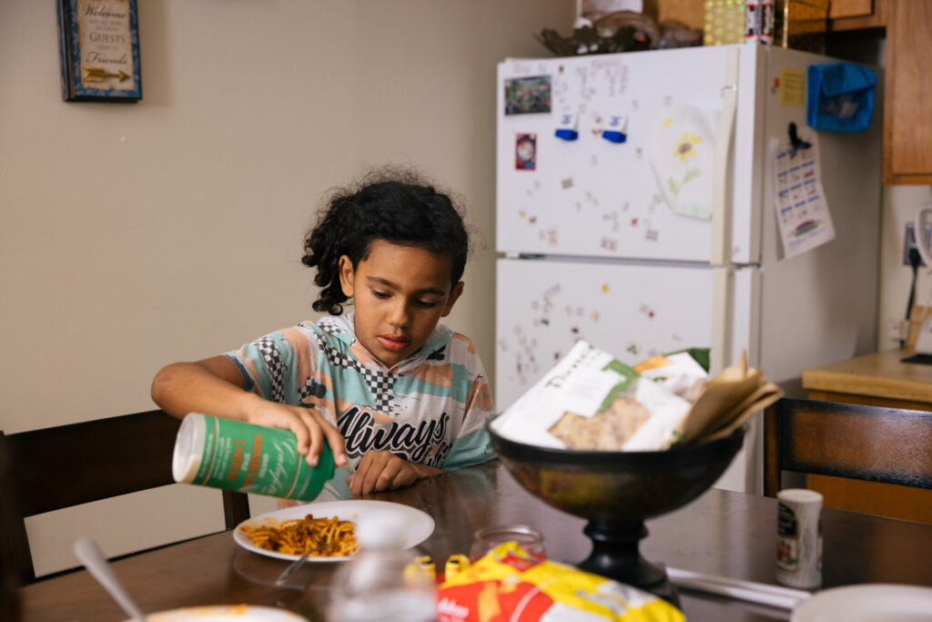 Drealyn sits at a kitchen table, pouring parmesan cheese onto his spaghetti.