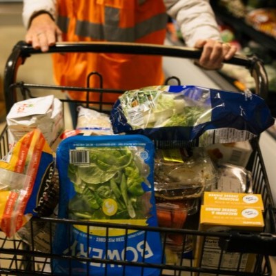 A person wearing a fluorescent orange vest pushes a shopping cart full of produce. 