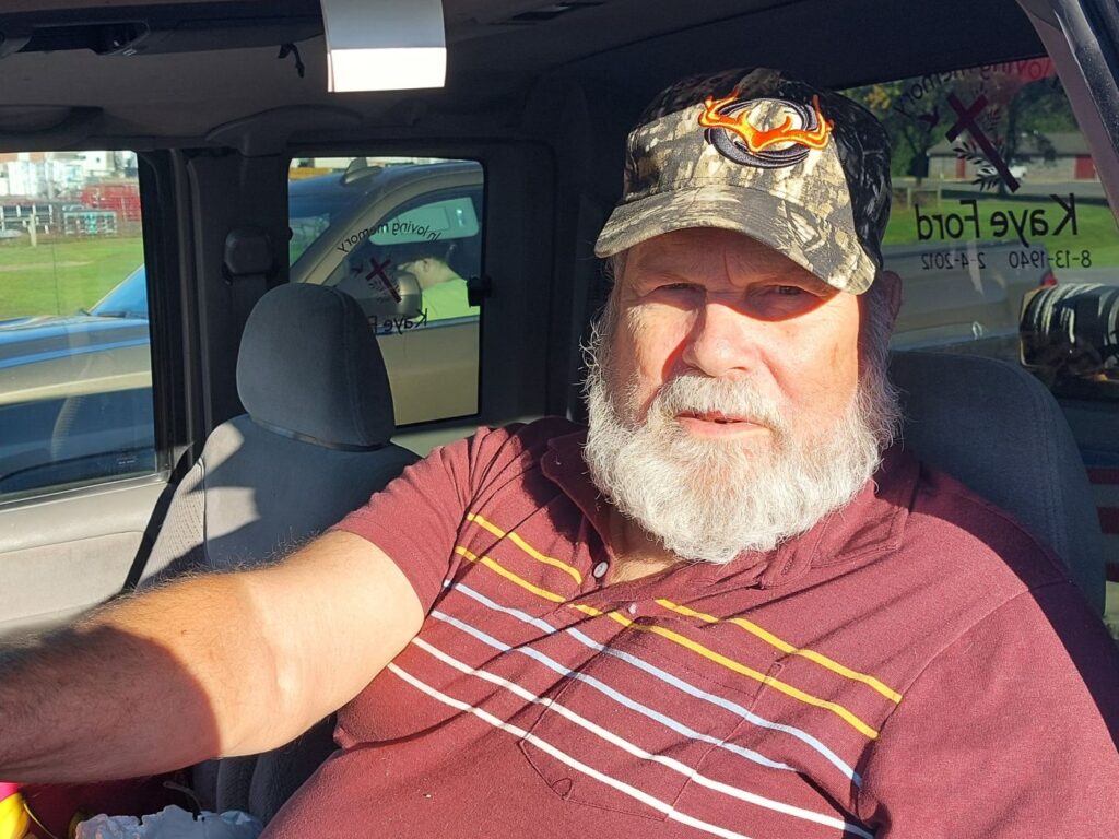 Berlin wears a red, striped shirt and a camo-printed hat. He looks at the camera from inside his truck with a serious expression on his face.