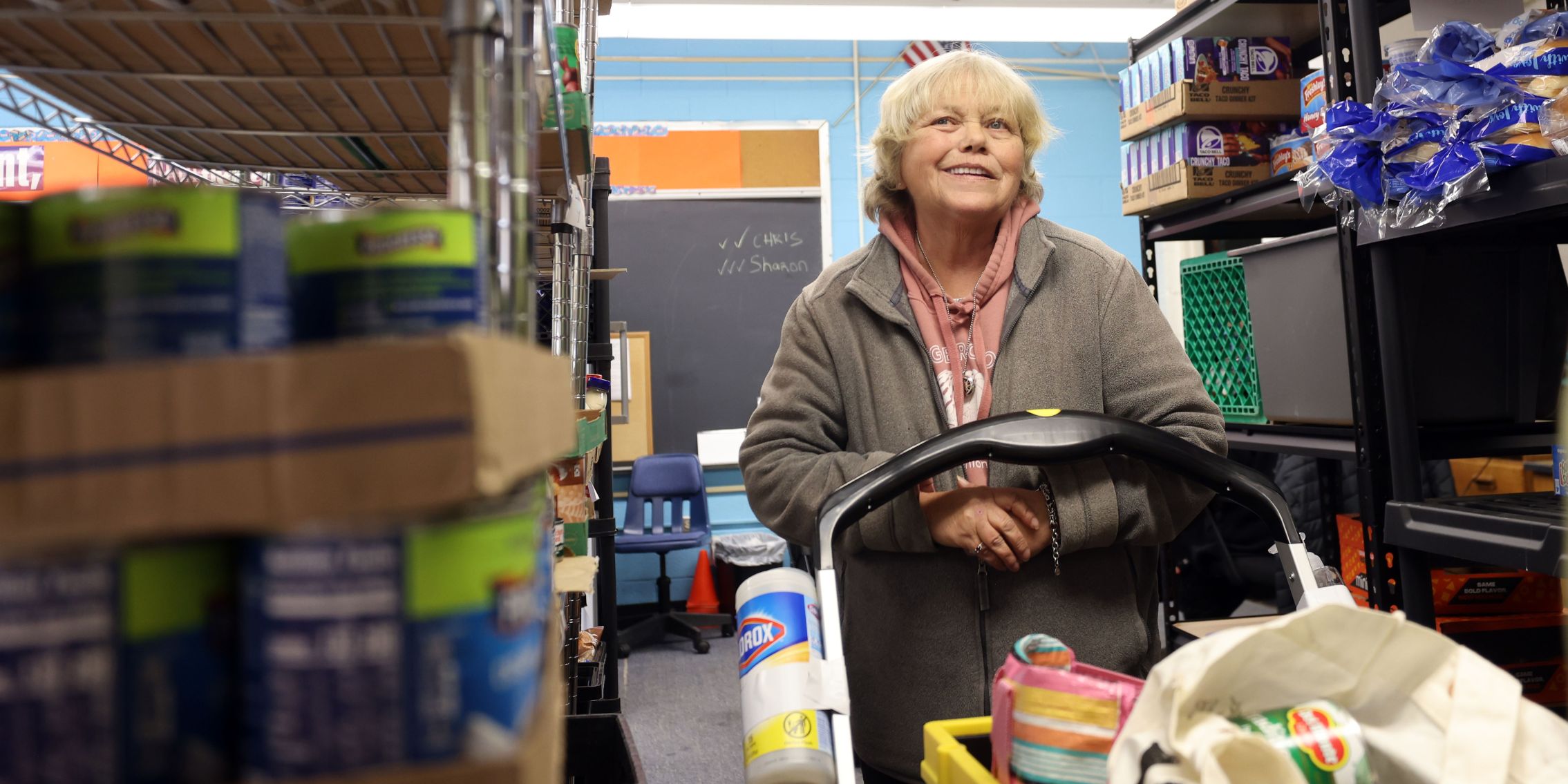 A guest browses the isles of a food pantry, pushing a shopping cart and smiling.