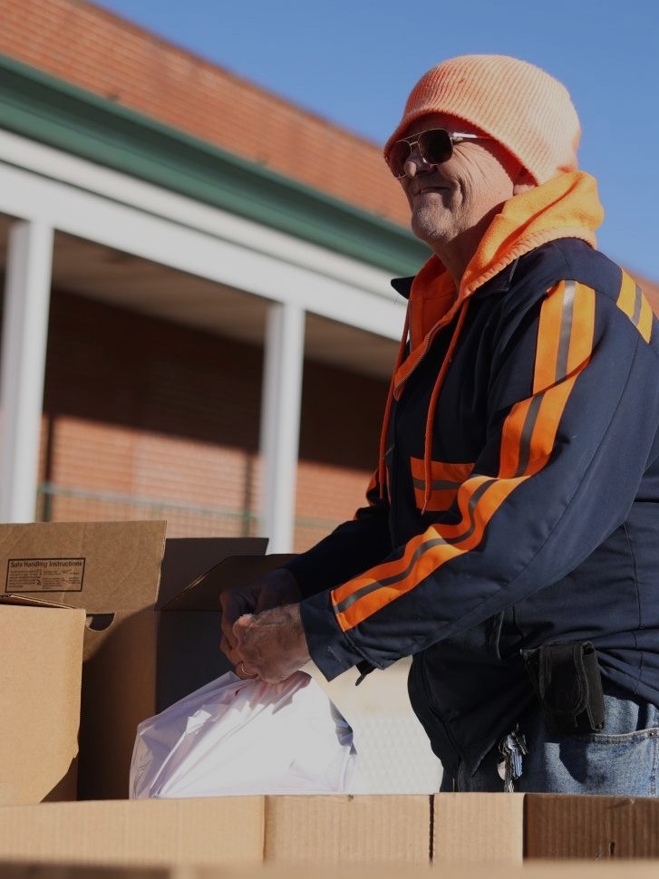 A Mobile Food Pantry Guest lifts a bag of food from a large cardboard box, smiling.